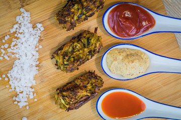 Homemade zucchini tots with condiments in decorative serving spoons and sea salt on wooden board top view