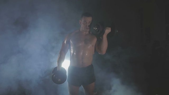 Strong sportsman lifting heavy dumbbells in smoke