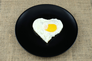 Fried egg in shape of heart on black plate.