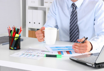 Cheerful young businessman sitting at the table with laptop and stretching in the office