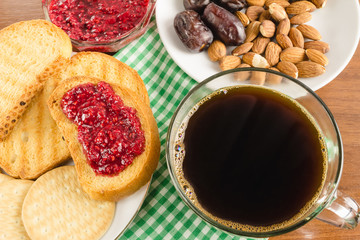 Breakfast time, cup of coffee with toasts, raspberry jam, dates, almonds at napkin on wooden table.