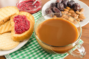 Breakfast time, cup of coffee with milk, toasts, raspberry jam, dates, almonds at napkin on wooden table.