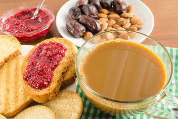 Breakfast time, cup of coffee with milk, toasts, raspberry jam, dates, almonds at napkin on wooden table.