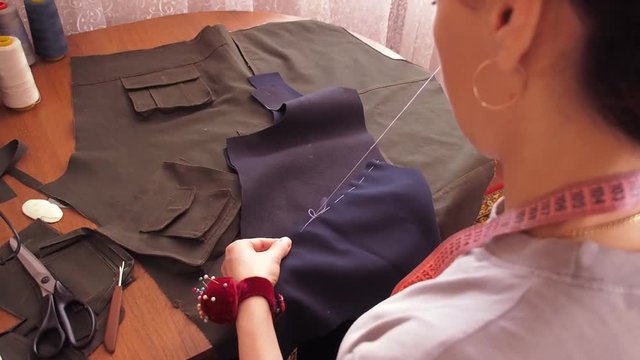 A seamstress sews by hand. Girl sits at the table and stabs baste fabric. Seamstress working at the table with a cloth. A woman in a gray T-shirt.