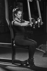 Fototapeta na wymiar Woman at the sport gym doing arms exercises on a machine. Dark sport club. Black and white.