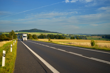 Fototapeta premium White truck arriving from a distance on the asphalt road between agricultural fields in a rural landscape