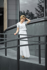 Fototapeta premium Blond hair girl in white dress stands on the metal stairs, on the gray background wall. Smiling and posing in front of a window.