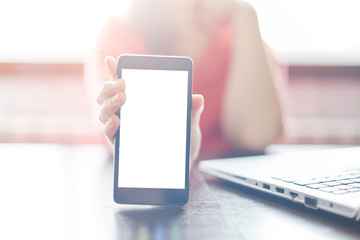 A cropped shot of female holding cell phone with blank screen for your promotional content. Girl showing something interesting on cell phone while sitting at table near her laptop.