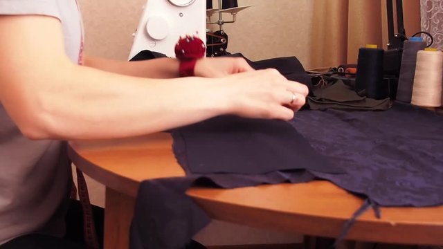 A seamstress sews by hand. Girl sits at the table and stabs baste fabric. Seamstress working at the table with a cloth. A woman in a gray T-shirt.
