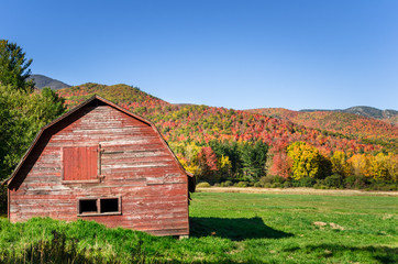 Obraz premium Disused Red Barn in a Grassy Field with Colourful Forested Mountains in Background