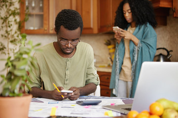 Naklejka premium Stressed African male in glasses holding mobile phone, using online banking app for paying gas and electricity bills, sitting at kitchen table with papers and laptop. Woman using gadget in background
