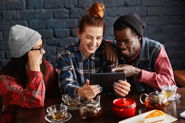 Three hipsters having fun at coffee shop: redhead woman with hair bun using touch pad with her African-American boyfriend and brunette girl while relaxing at cafe, drinking tea, watching videos online