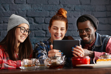 People, international friendship, leisure, modern technology and communication concept. Three students having rest indoors, enjoying wireless internet connection at cafe and using electronic tablet