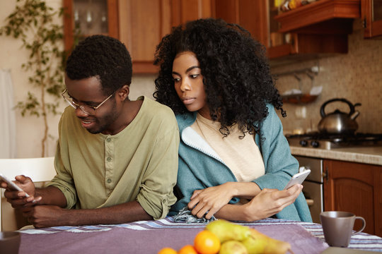 Portrait of sneaky and jealous young African wife spying on her husband, looking over shoulder, trying to see what he reading on his mobile phone. People, relationships and modern technology