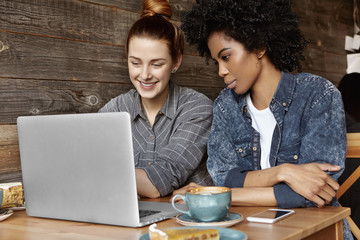 Two stylish lesbians sitting at cafe in front of open laptop, surfing internet, watching videos online during coffee break. Caucasian girl showing something on notebook pc to her African girlfriend