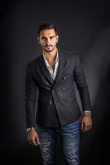 Obraz premium Young businessman confidently posing in front of camera, wearing business suit without neck-tie, with shirt open on neck, on dark background