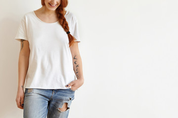 Fototapeta premium Cropped shot of beautiful stylish young redhead woman with braid and tattoos smiling joyfully dressed in white oversize t-shirt, keeping hand in pocket of her trendy blue ragged jeans. Horizontal