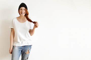 Hipster girl wearing black stylish hat, white t-shirt and ragged jeans smiling cheerfully, touching her ginger hair in braid, posing against studio wall with copy space for your promotional content