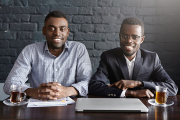 Portrait of two handsome confident African-American recruiters sitting close to each other at desk with papers and generic laptop pc, looking and smiling at camera, waiting for next job candidate
