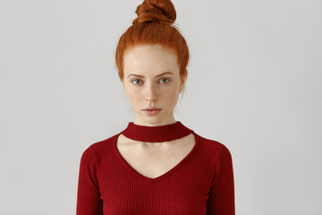 Beauty, style, fashion, clothing and design concept. Gorgeous luxurious Caucasian redhead woman with hair bun and freckles wearing stylish red dress with geometrical cut out before going out on date - obrazy, fototapety, plakaty