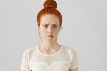Confused or puzzled beautiful young Caucasian woman with ginger hair frowning, biting her lower lip after having done something wrong, looking at camera with guilty and apologetic facial expression - obrazy, fototapety, plakaty