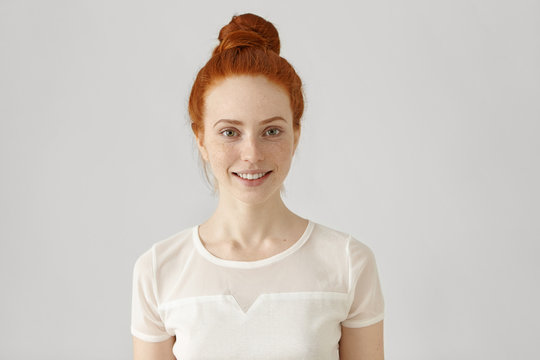 Cheerful young Caucasian woman with freckles and ginger hair in bun looking and smiling happily at camera, wearing white blouse. Redhead girl with pretty joyful smile posing at blank studio wall