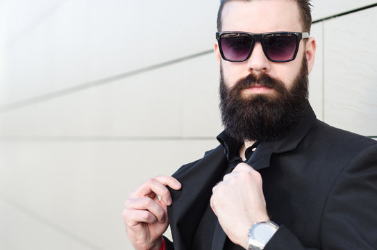 amazing portrait of a man with a long beard and glasses in a black suit, serious hipster man looks into the camera
