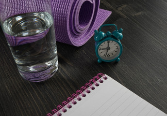 Healthy lifestyle concept - glass of water, yoga mat, alarm clock and notebook on the dark wooden background