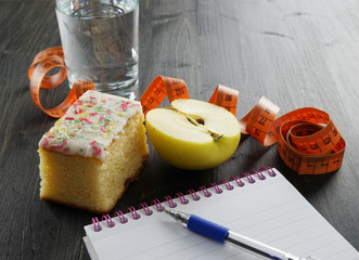 Healthy lifestyle concept - glass of water, apple, cake and notebook on the dark wooden background, weight loss concept