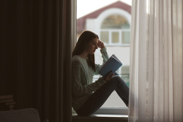Fototapeta premium Young woman reading book on windowsill at home