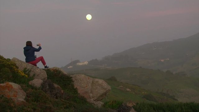 Girl taking photos selfie on smartphone, while sitting on a rock hill, beautiful evening moon in the mountains, nature sunset