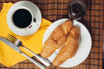 croissants in a plate, a cup with a drink and a fork with a knife on a yellow napkin