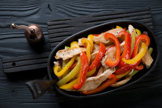 Cast-iron frying pan with chicken fajitas on a black wooden serving board, high angle view