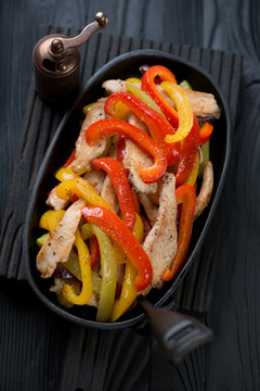 Tex-mex chicken fajitas served in a cast-iron frying pan on a black wooden background, studio shot