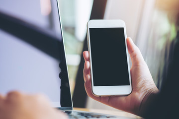 Mockup image of a hand holding white mobile phone and blank black screen with laptop in modern cafe