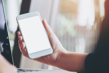 Mockup image of a hand holding white mobile phone and blank white screen with laptop in modern cafe