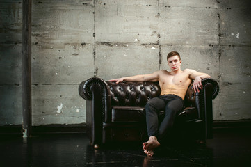 Fototapeta na wymiar Young sexy men bodybuilder athlete sitting on a black leather couch with a naked torso and black trousers, studio portrait