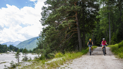 Fototapeta na wymiar Mountain biking woman and young girl along river in Dolomites, Italy