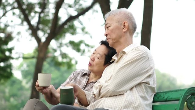 Slow motion happy Asian senior couple sitting and drinking tea, coffee in morning park. Having conversation and showing love