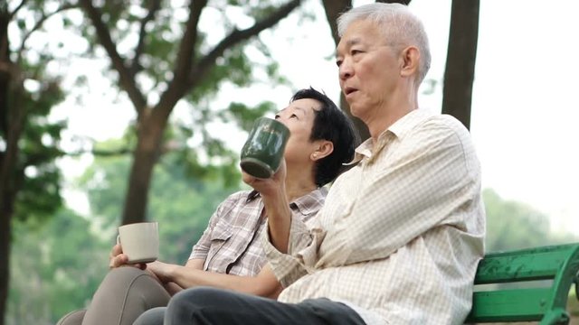 Slow motion happy Asian senior couple sitting and drinking tea, coffee in morning park. Having conversation and showing love