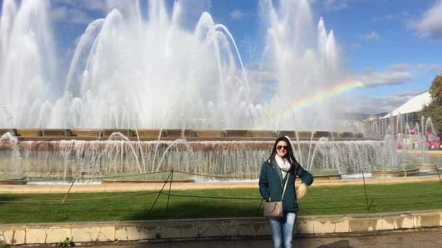 Young woman posing to camera by large funtain and natural rainbow. Girl waving her hand to camera posing for photo