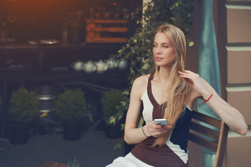 Portrait of blonde pensive beautiful girl using smart phone in street cafe, charming caucasian...