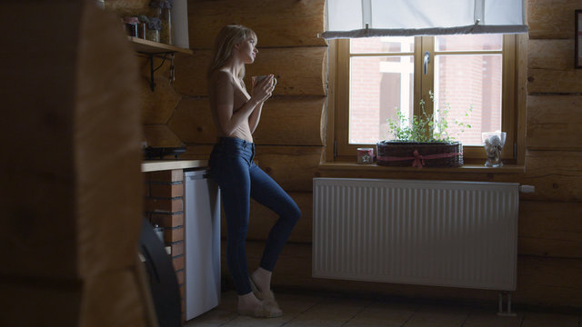 Beautiful young pensive woman holding cup of hot tea and standing by the window. 