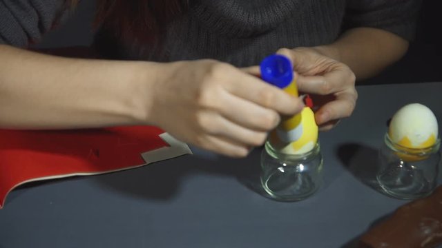 Girl in a dark sweater decorates yellow egg sticking handmade comb and beak from red paper.Creating easter chicken from the egg shell by hand on a grey table close up.