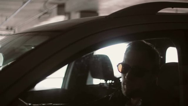 Young Smiling Man In Sunglasses Driving Car. Male Sitting Inside, Reverse And Opens The Car Window At The Parking Place.