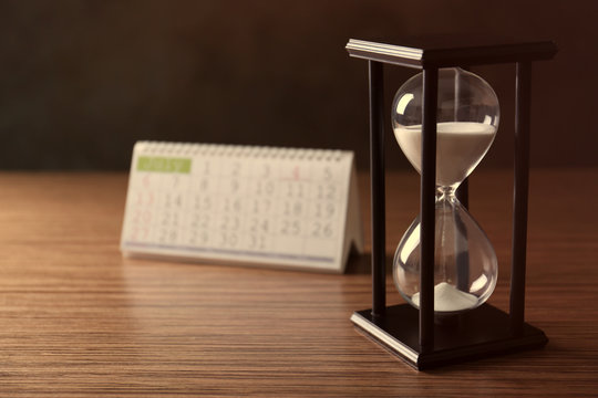 Black hourglass with white sand and calendar on wooden background