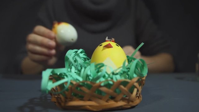 Women's hands are placed in small wicker openwork bamboo basket with green paper grass two yellow easter chicks of eggshells handmade over a gray background close up.