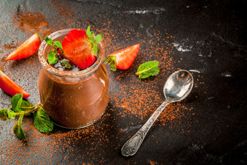 Chocolate dessert (yogurt, pudding) in a jar for one serving, dusted with cocoa with mint leaves and fresh strawberries. On a black table, copy space