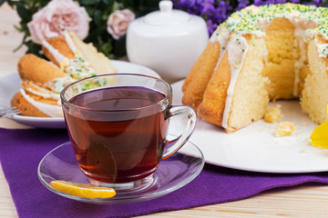 Homemade cake with icing sugar sprinkles on a plate and cup of tea on a wooden table with purple flowers and pink roses in the background