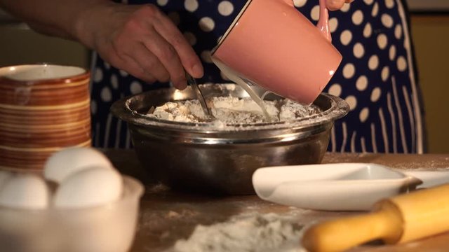 Close up shot of baker's hands who kneads the flour spoon, pouring water into the flour out of the cup. To prepare the dough also needed eggs, which lie in a bowl on a table.